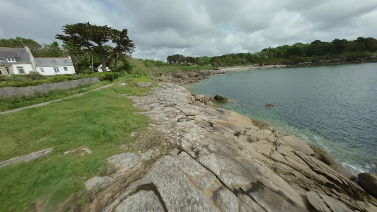 FPV drone flies closely above large sea rocks, with lush trees lining shoreline under cloudy skies in Concarneau, Brittany, France, natural coastal beauty.