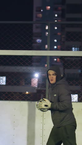 el portero lanza una pelota de fútbol durante la práctica nocturna cerca del poste de la portería con la sombra proyectada en la pared detrás, enmarcada por edificios residenciales iluminados en el fondo urbano