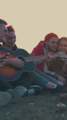 group of young people sit at burning bonfire and guy plays guitar in camp on meadow at evening sunlight