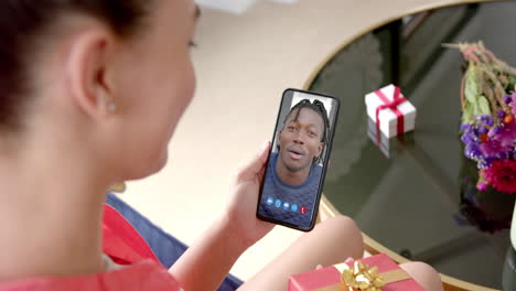 Biracial-woman-holding-smartphone-with-african-american-manon-screen-with-gift-on-desk