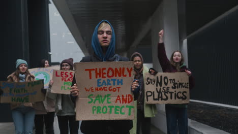 young male activist holding a cardboard placard during a climate change protest while looking at camera surrounded by others activists