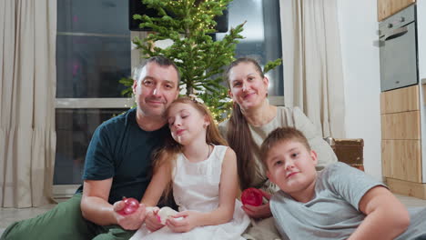 father mother and two kids sitting on soft rug in cozy modern kitchen holding colorful christmas ornaments in hands smiling happily together near decorated tree during festive winter holiday