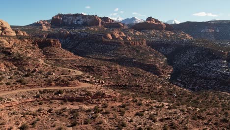 Eine-Hochfliegende-Drohne-Schoss-über-Eine-Abgelegene-Unbefestigte-Straße,-Die-Durch-Das-Weite-Und-Einzigartige-Wüstenland-In-Der-Nähe-Von-Moab,-Utah,-Führt,-Mit-Den-Schneebedeckten-Rocky-Mountains-In-Der-Ferne