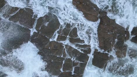 Top-view-moving-up-of-sea-foam-crashing-rocks