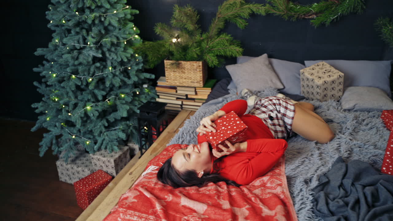 Pretty girl in cozy bedroom at Christmas. Beautiful young woman lying on bed with a gift in New year night. Happy girl at home in festive decorated room.