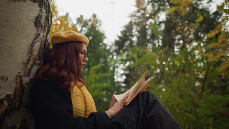 college student sits in solitude leaning against tree, deeply engaged in book, flipping pages thoughtfully in peaceful outdoor setting surrounded by autumn leaves and nature beauty