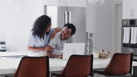 couple wearing pyjamas standing in kitchen working from home on laptop