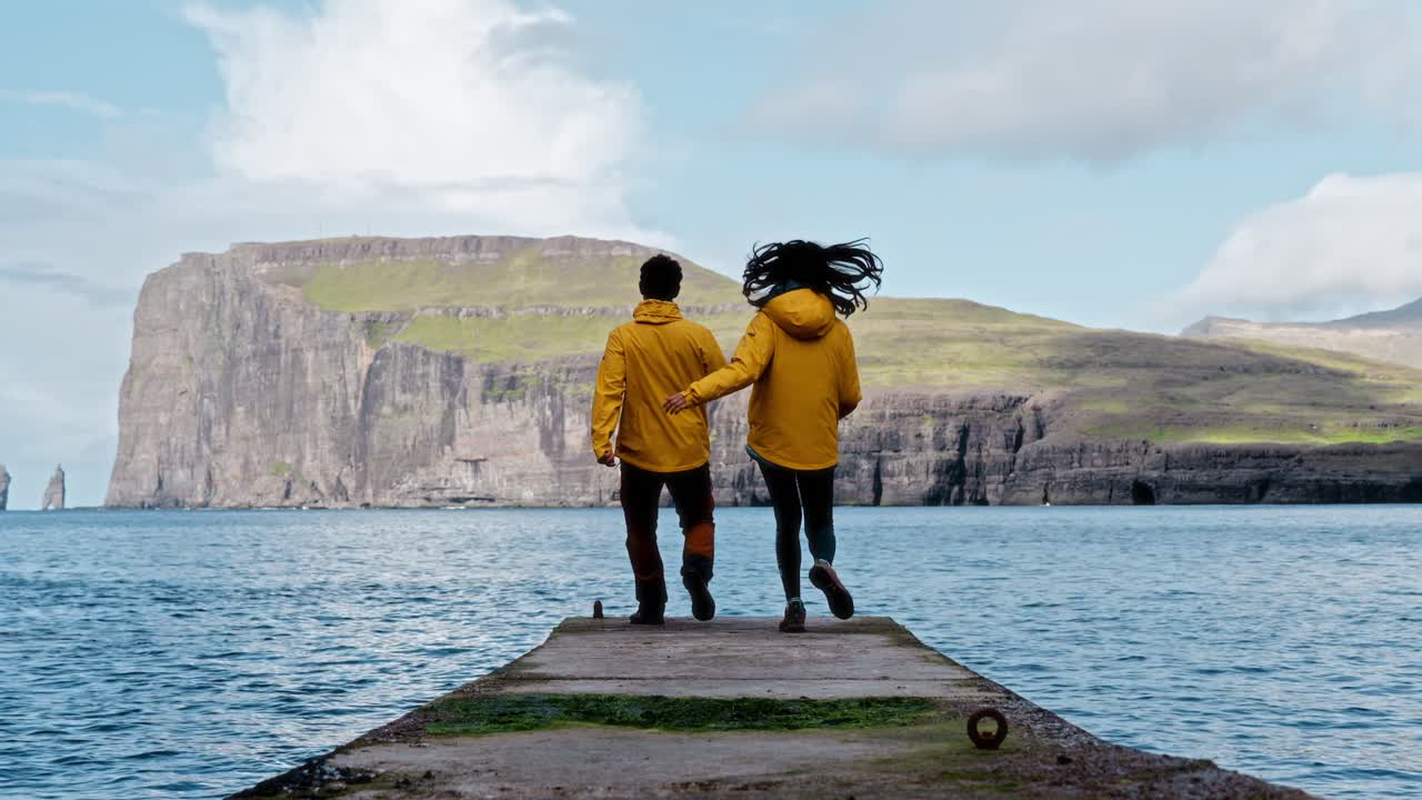 Two people in yellow jackets run hand in hand towards a dock, with scenic cliffs and ocean views in the background, evoking freedom and joy