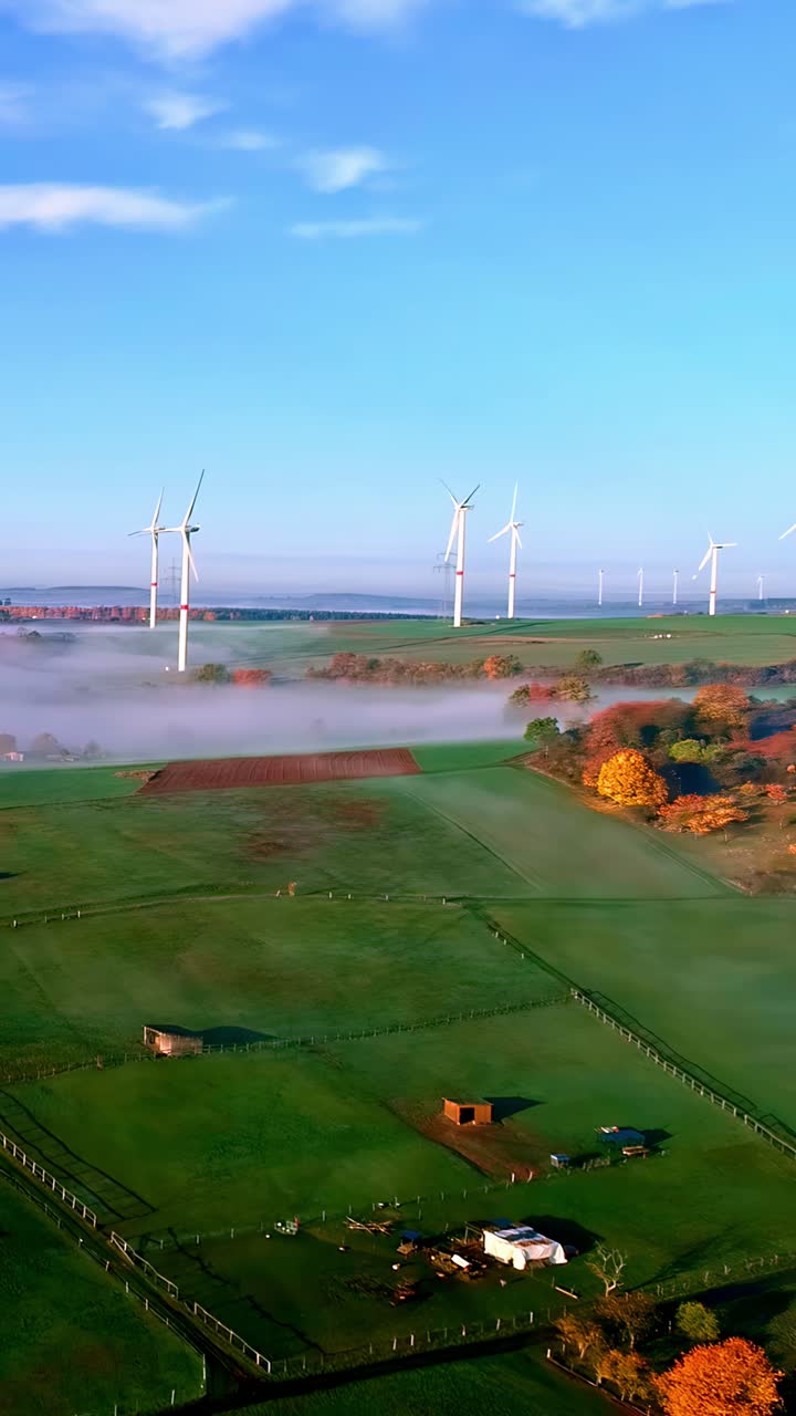 Drone flies toward wind turbines spinning above misty farmland and rural fields at sunrise