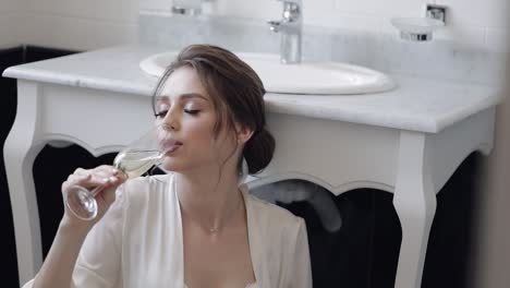 Bride-in-white-boudoir-dress-sitting-on-floor-in-bathroom-at-home-apartment-with-glass-of-champagne