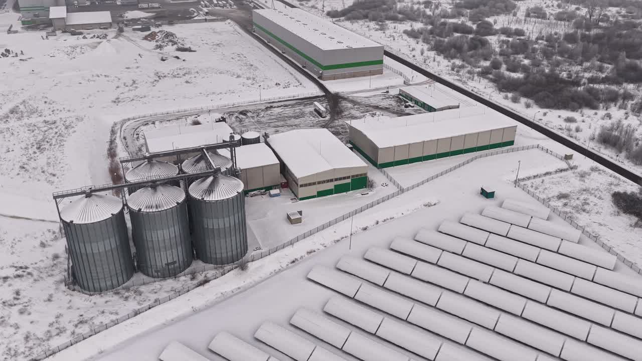 Drone aerial of snow-covered grain storage facility with metal silos, warehouses, and logistics yard in rural landscape, cold season industrial agriculture complex