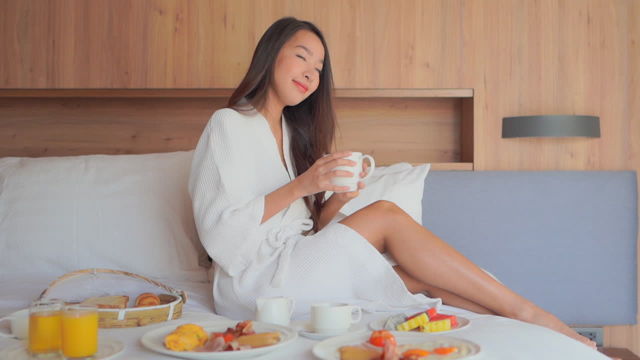 하얀 목욕 가운을 입은 아시아 여성은 침대에서 아침을 먹고, 커피 한 잔을 마시고 홀짝인다