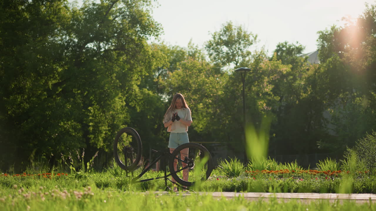 젊은 여자가 거꾸로 된 자전거 에 기게 서서 팔을 들고 자전거를 보고 흥분해서 뛰어내립니다. 태양빛은 배경에서 따뜻하고 밝은 효과를 만니다.