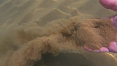 Male-hand-grabs-sand-from-seabed-and-slowly-lets-it-flow-through-fingers,-underwater-slow-motion-view