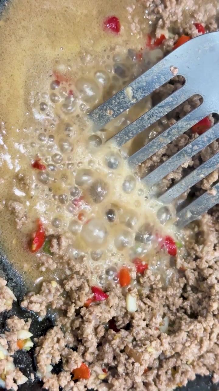 Hackfleisch, rote Chili und Currysauce sprudeln kräftig in einer Bratpfanne unter heller Beleuchtung, wobei ein Metallspatel die Mischung von oben umrührt