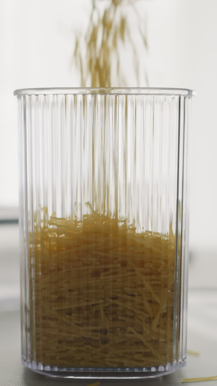 Broken pasta falls into tall plastic container on table. Person pours raw product into packing for storage in light kitchen. Maintaining order in house