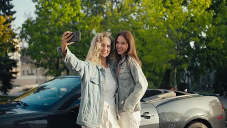 2 chicas felices rubia y morena se toman una selfie en el fondo de un cabriolet gris en verano