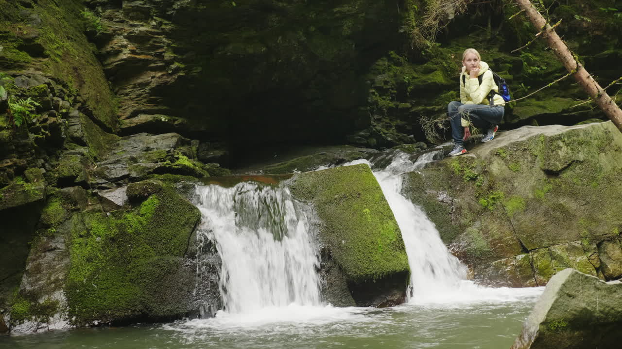 숲 4k 비디오에서 폭포를 감상하는 노란색 다운 재킷을 입은 여성