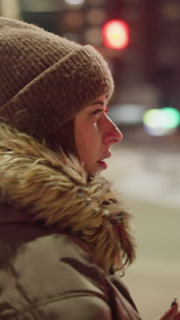 first-person view of a woman in a winter coat with a fur hood, standing and rubbing her hands together while looking to the right at night. blurry cars are moving in the background