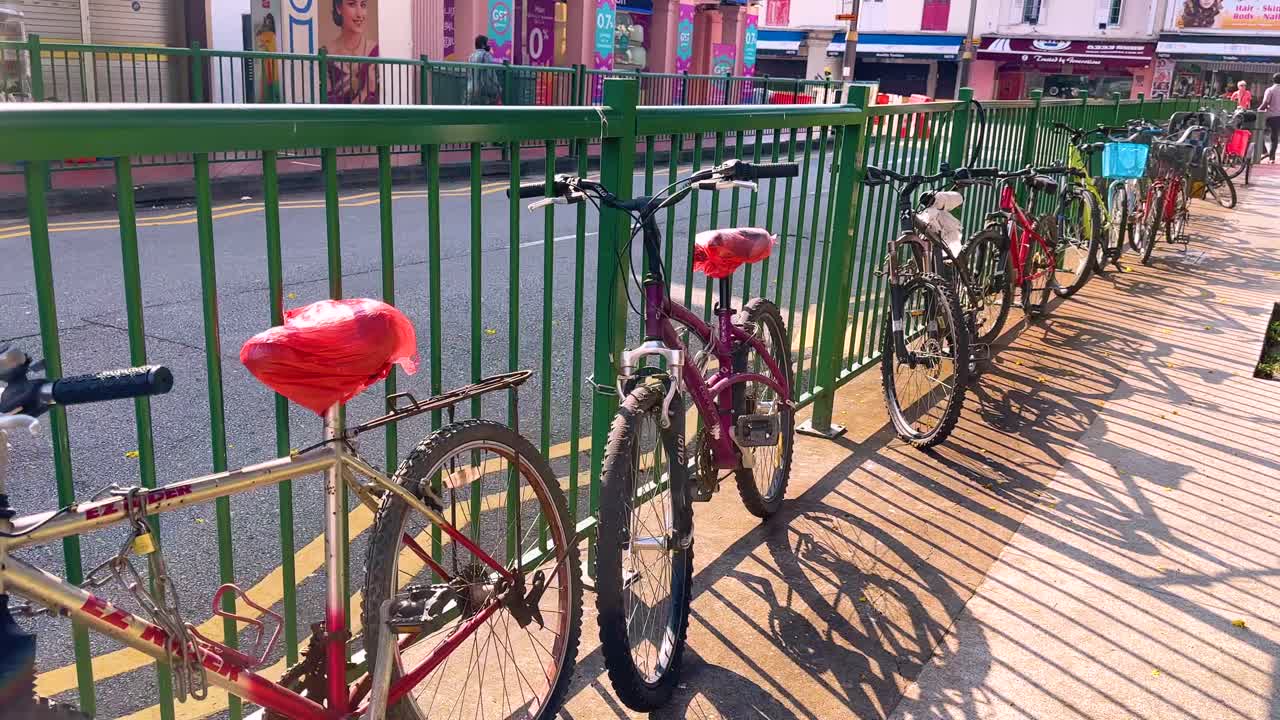 Multiple bicycles are parked and locked to a green railing on a sunlit sidewalk in Singapore, captured with smooth lateral camera movement and strong midday shadows
