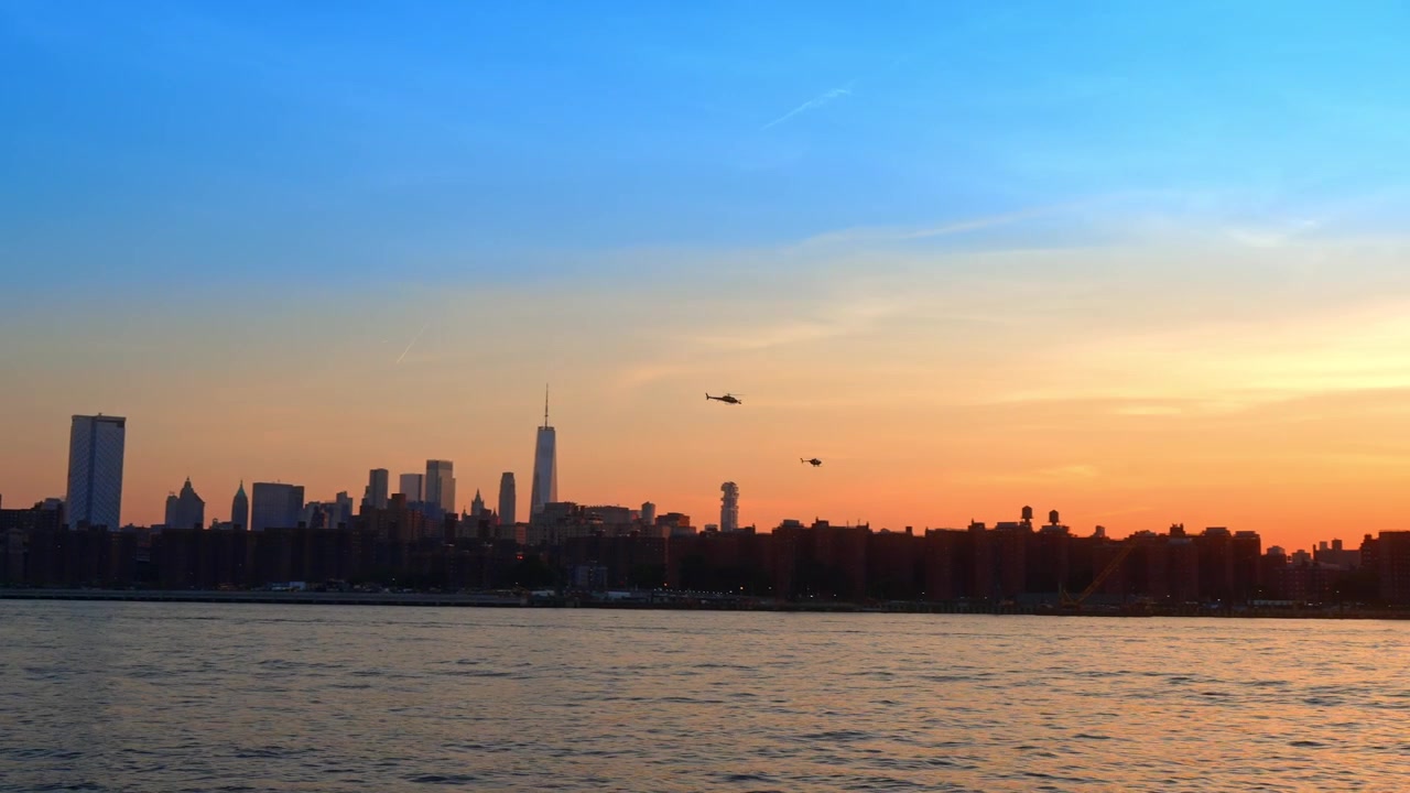 Helicopter flying over lower Manhattan skyline at sunset, New York. Distant aircraft silhouette with Williamsburg Bridge, pier, and gradient sky from Brooklyn.