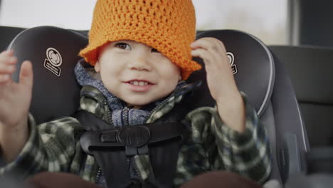 un alegre niño asiático viaja en un asiento de coche para niños, divirtiéndose tirando de un sombrero sobre su cabeza. un paseo divertido