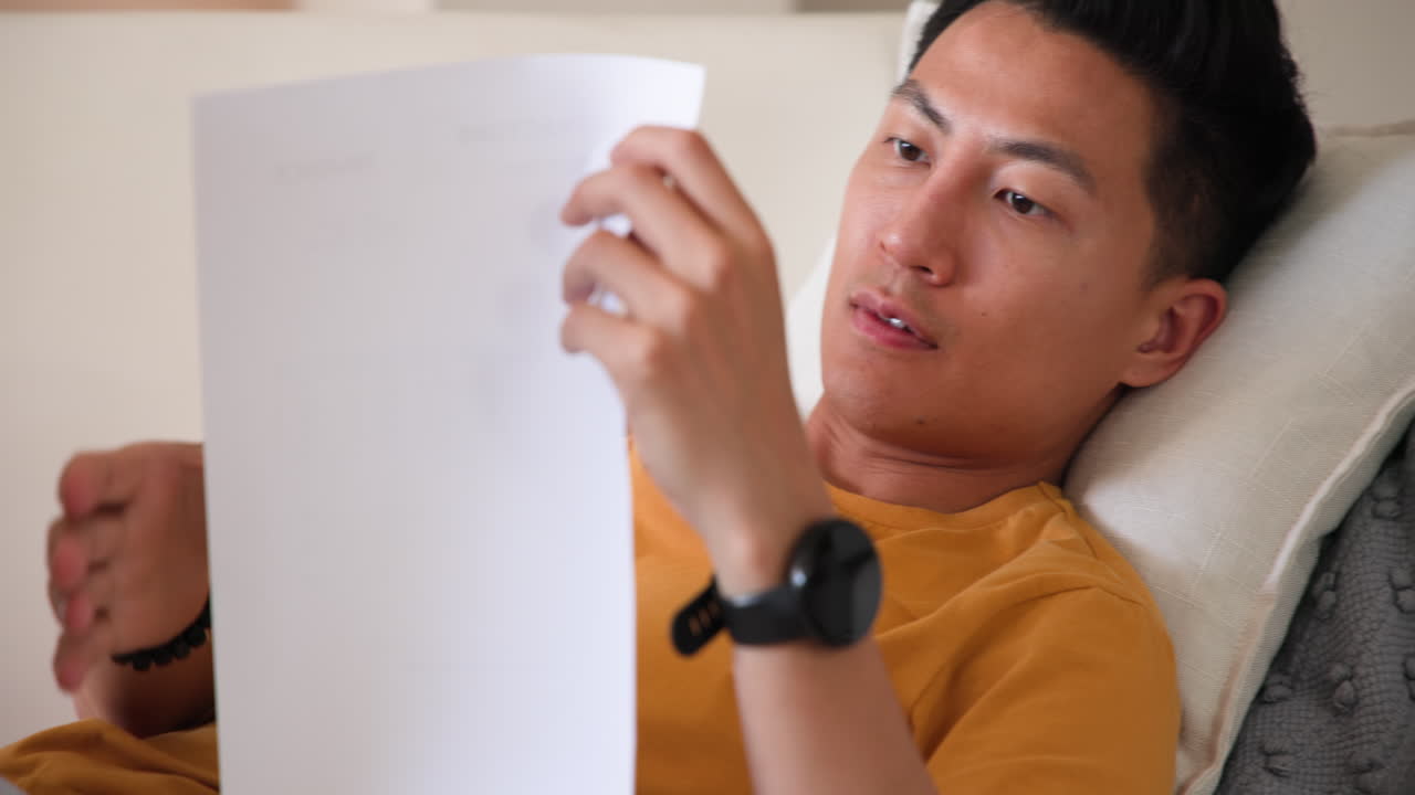 Relaxing at home, man reading document while lying on couch, wearing watch