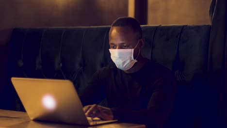 male typing on a laptop in a dark café wearing a facemask