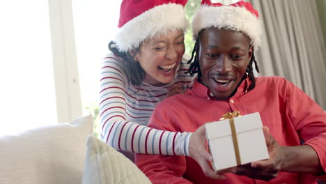 happy diverse couple in christmas hats exchanging gift in sunny living room, slow motion