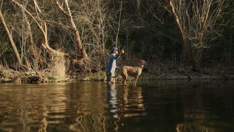 Pescador-Masculino-Y-Su-Perro-Se-Paran-En-Medio-Del-Río-Durante-La-Hora-Dorada-Lanzando-Caña-De-Pescar-Al-Atardecer