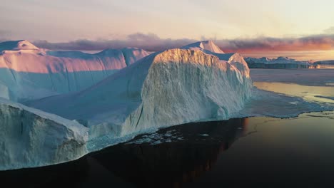 Vista-Aérea-De-Icebergs-Iluminados-Por-El-Sol-Y-Reflejo-Del-Mar-En-Calma,-Espectacular-Puesta-De-Sol-En-Groenlandia