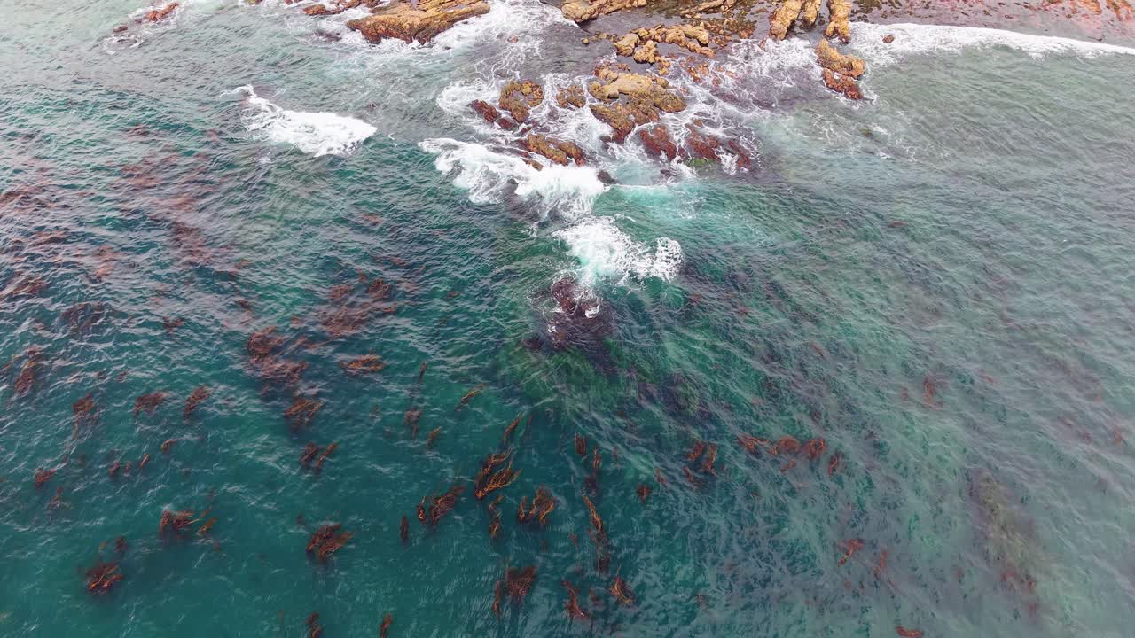 Drone shot of ocean surf breaking against jagged rocks in shallow coastal waters