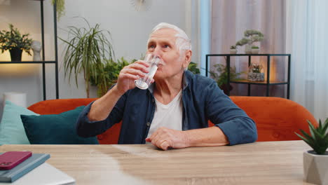 Thirsty-senior-old-man-sits-at-home-holding-glass-of-natural-aqua-make-sips-drinking-filtered-water