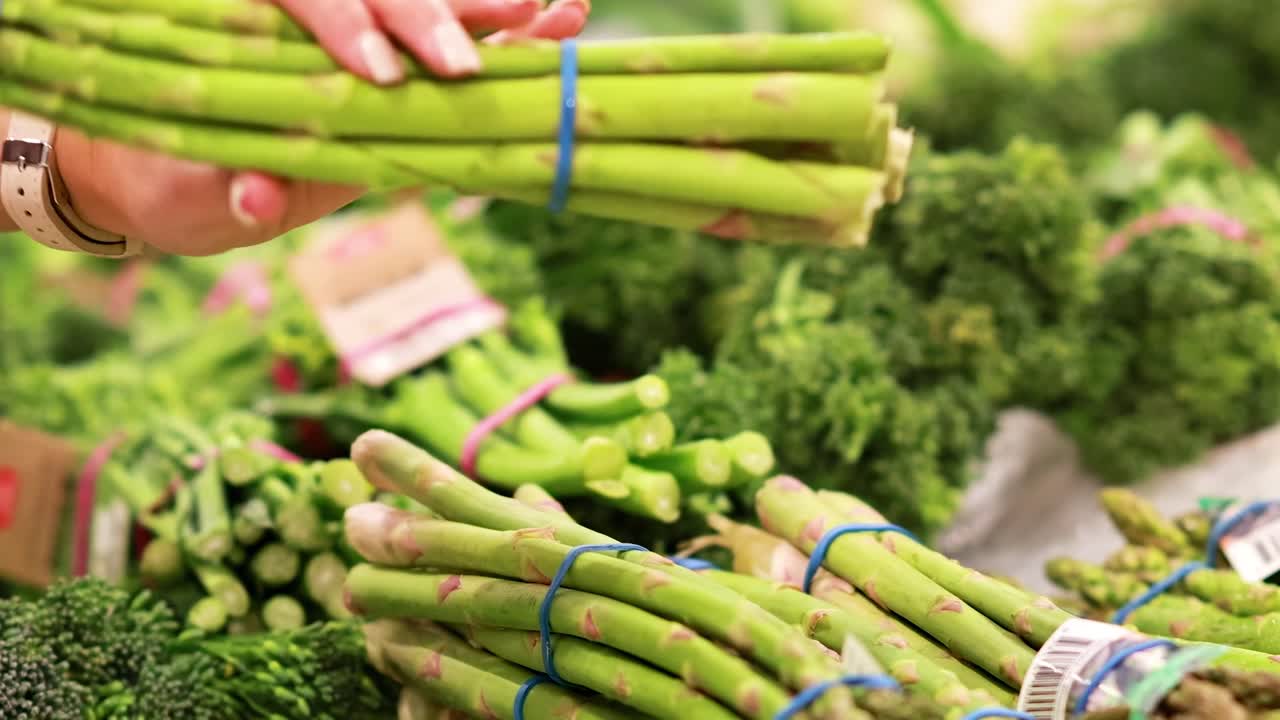 A person examines asparagus bundles in a well-lit supermarket produce section, focusing on freshness and quality