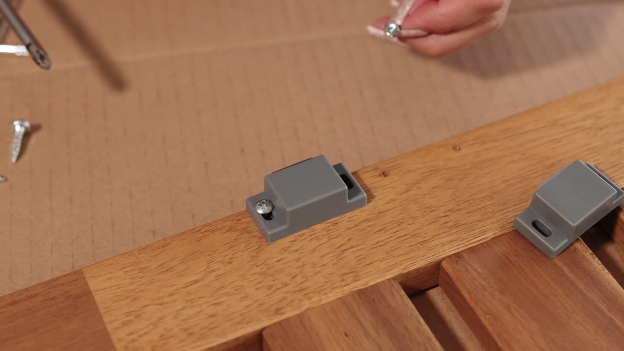 Hands assemble a magnetic catch on wooden furniture using screws. Close-up shots highlight precision and technique in a well-lit setting