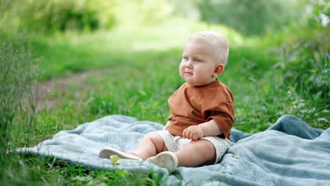 lovely blond baby boy in brown t-shirt and white shorts sits outdoors on the grass. cute toddler plays with a green leaf peacefully.