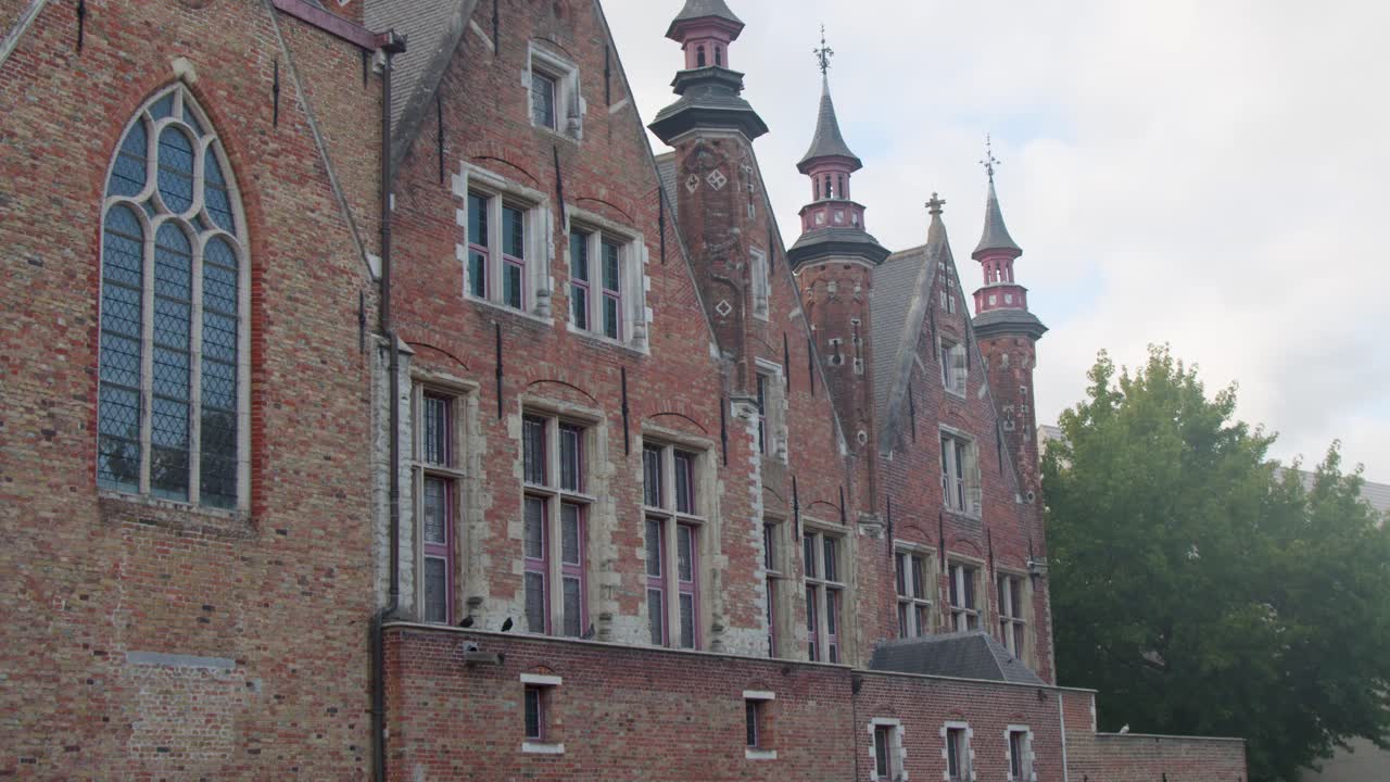 Camera slowly pans right, revealing historic red brick Gothic architecture beside tranquil canal in daylight