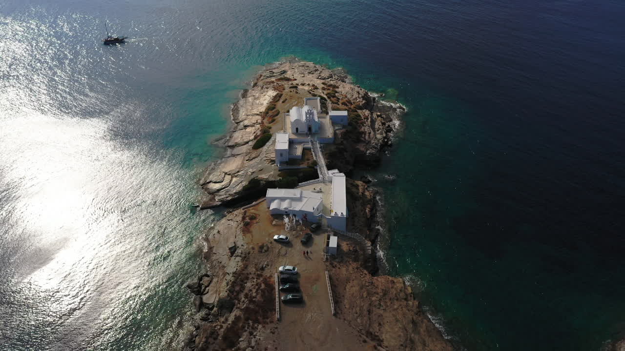 Aerial shot descending towards the church of Chrisopigi on the cycladic island of Sifnos, Greece. A small sailing boat sails by towards the suns reflection.