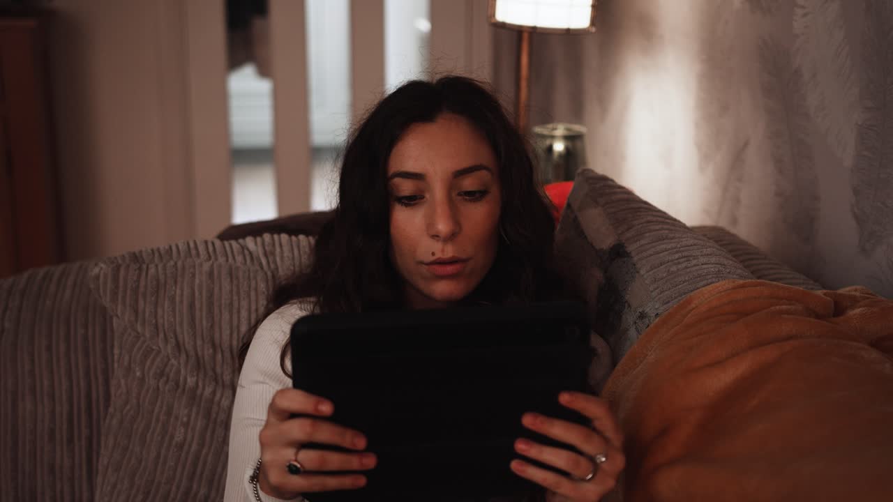 Young Italian woman smiles while relaxing using a tablet on the sofa