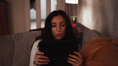 young italian woman smiles while relaxing using a tablet on the sofa