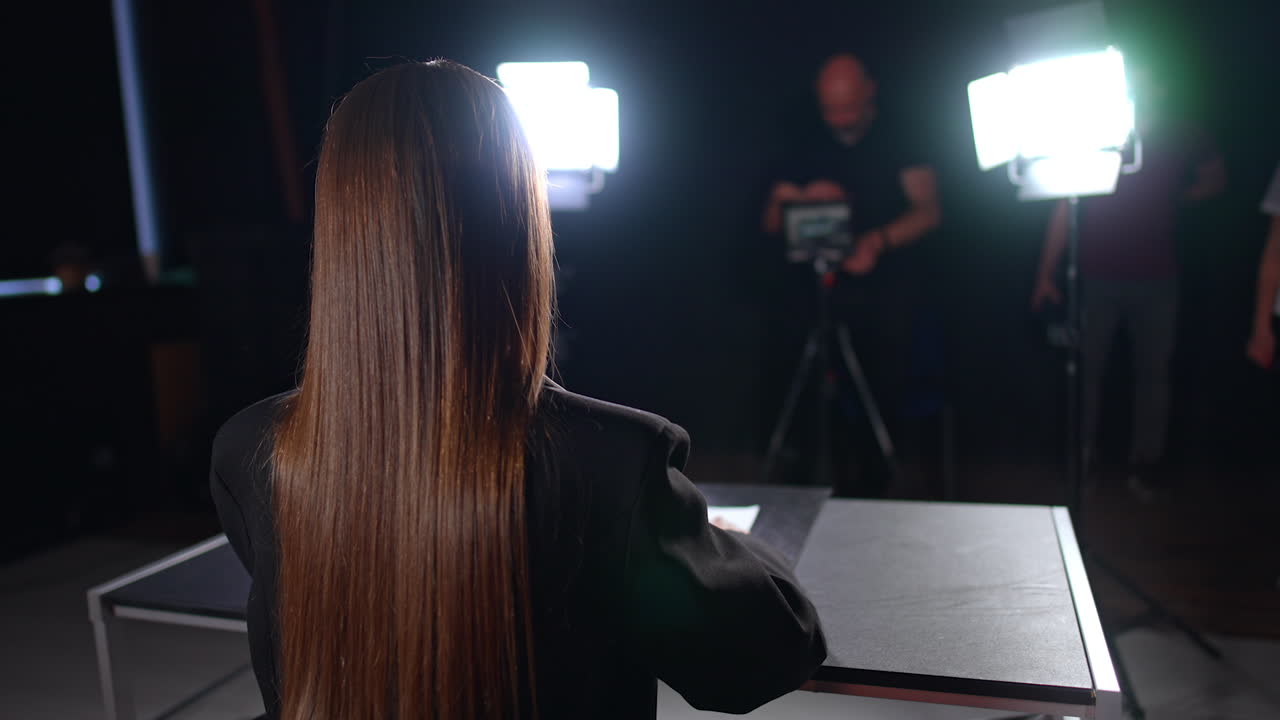 Rear view of a long-haired brunette woman in black jacket sits in the light of soffits. Cameraman taking video on professional camera at blurred backdrop.
