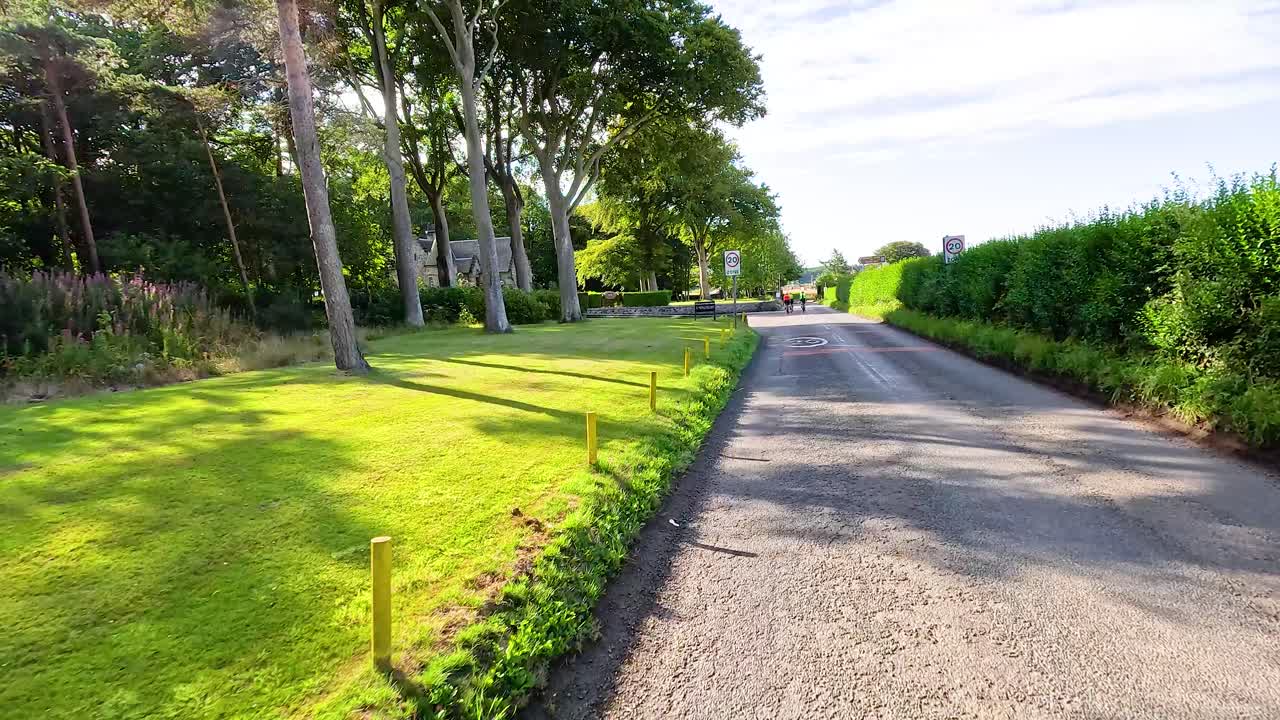 POV cycling down sunlit village road, stone walls, hedges, trees, and houses, smooth camera movement