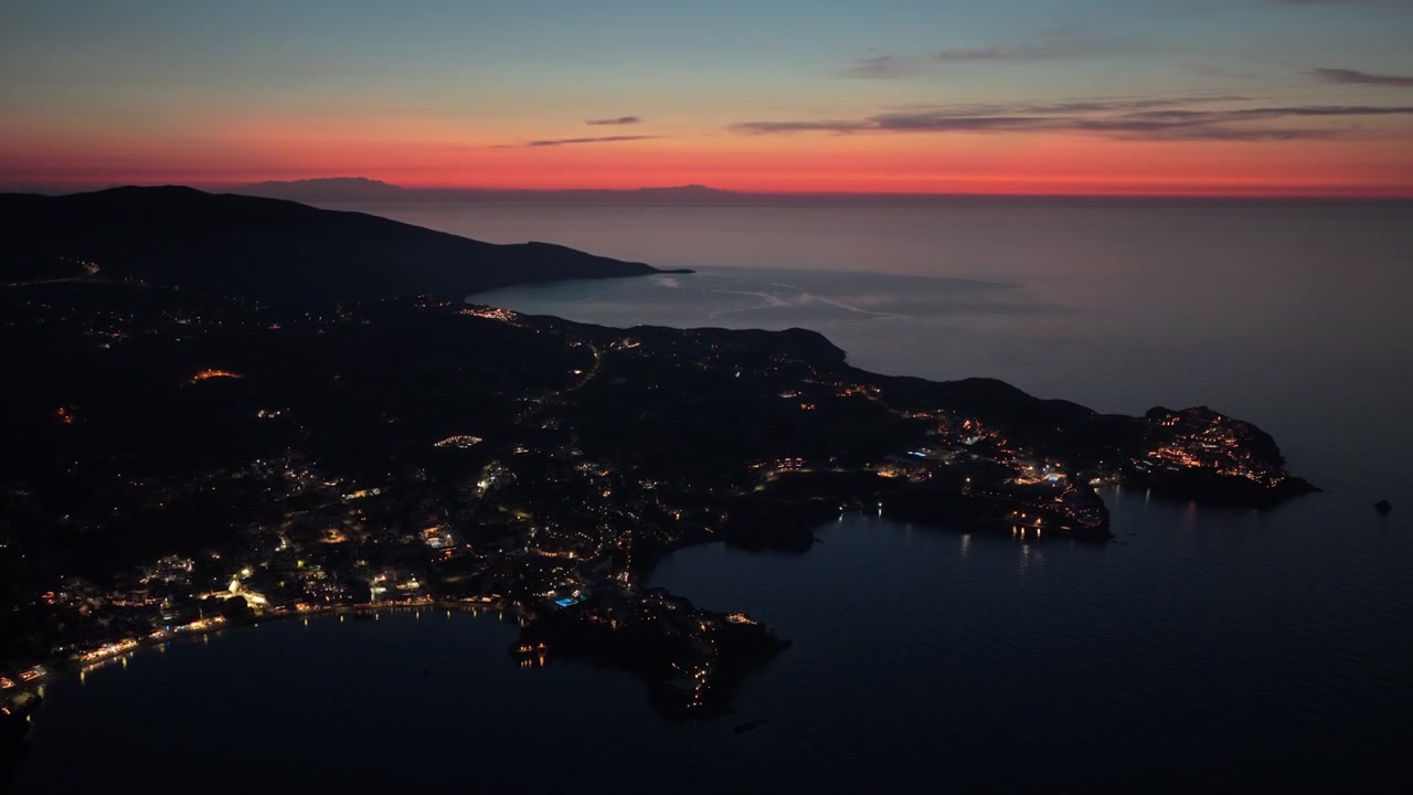 Høytliggende dronebilde av kystlinjen og nesene med spredte bylys under et rødt etterskin, Agia Pelagia, Hellas