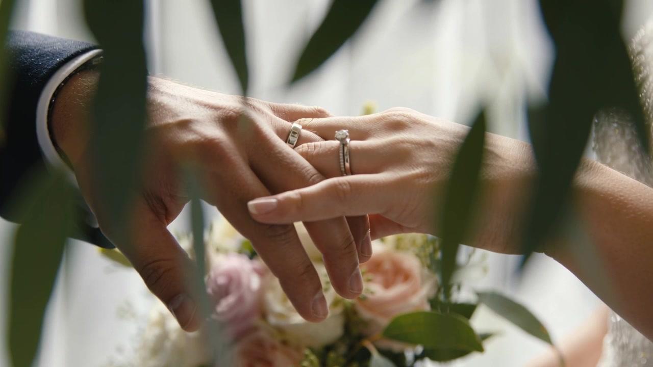 Hands approaching, groom's hand gently adjusting bride's ring at wedding, with bouquet, watch
