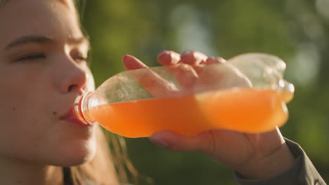 close-up of woman drinking orange juice outdoors from a plastic bottle with relaxed satisfaction, sunlight reflecting on the bottle and her hand, soft glow of light and greenery in the background