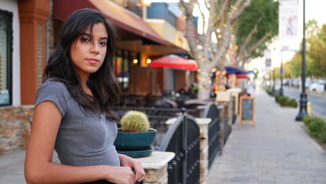 A-beautiful-hispanic-woman-standing-on-the-downtown-urban-city-street-full-of-shops,-restaurants-and-retail-stores-SLOW-MOTION