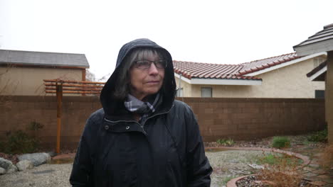 an elderly woman with glasses and a raincoat standing in a winter weather rain storm as raindrops fall in slow motion