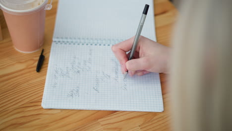 close-up of hand writing with black pen in a notebook on a wooden table, pen cover lies nearby, and a coffee cup is partially visible, creating a cozy workspace for note-taking and journaling