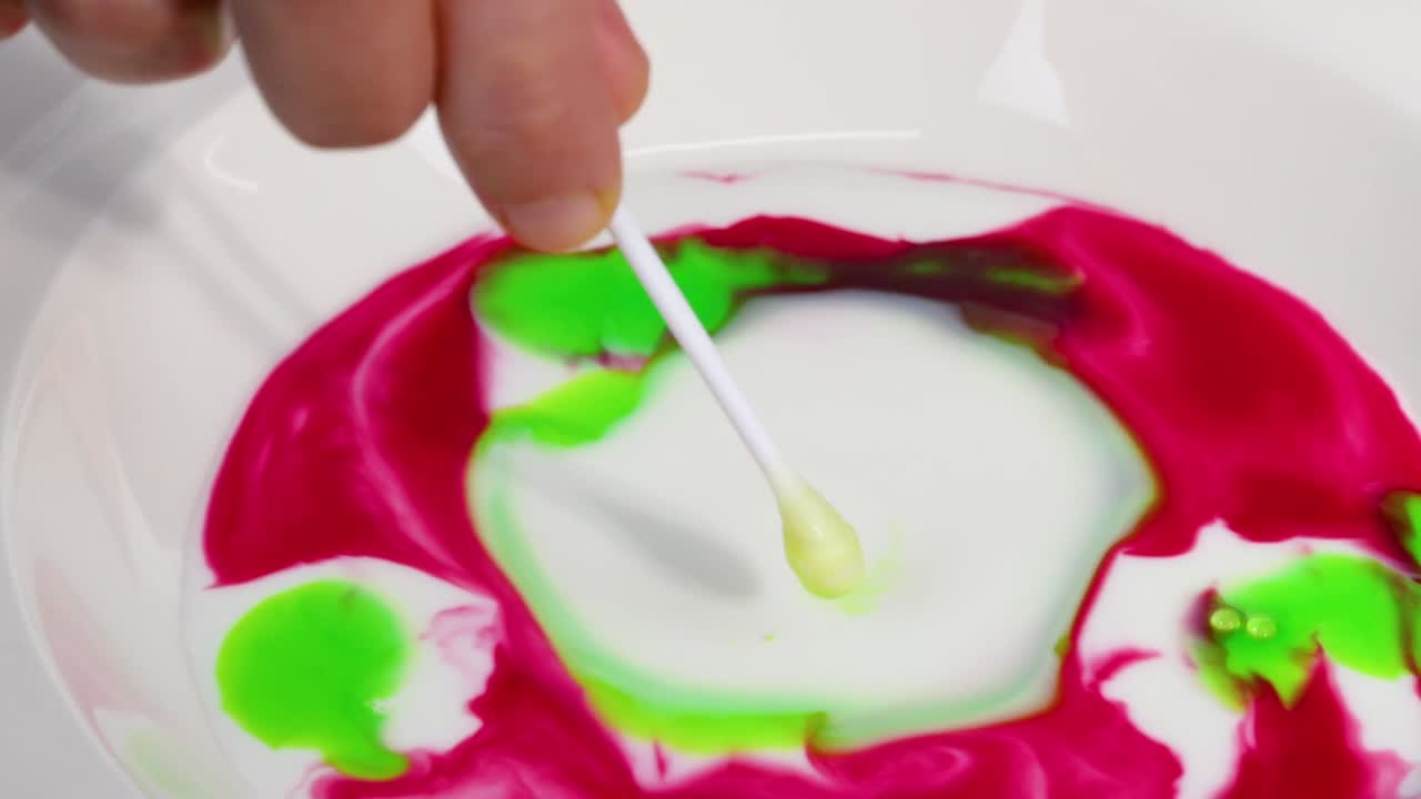 Observe the interaction of vibrant pink and green colors swirling dynamically in milk with a cotton swab.