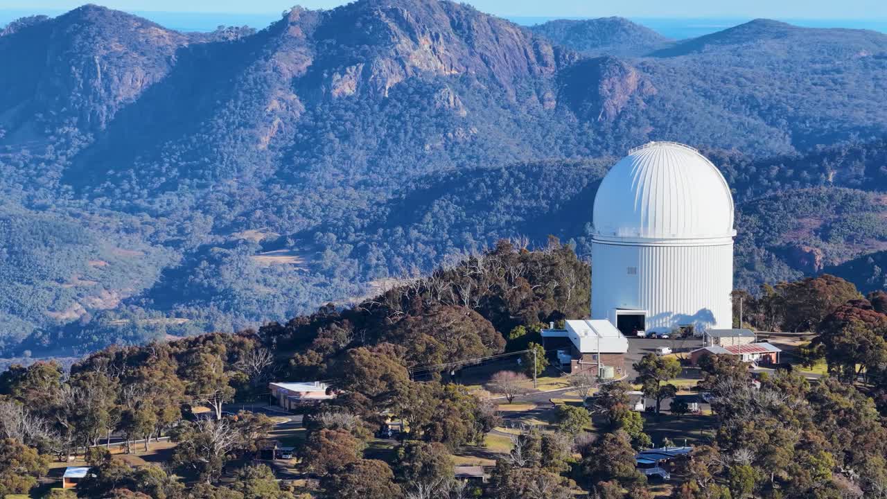 Aerial drone footage circles a large white astronomical observatory dome atop a forested ridge, with rugged mountain landscape and soft sunset lighting in Coonabarabran, New South Wales